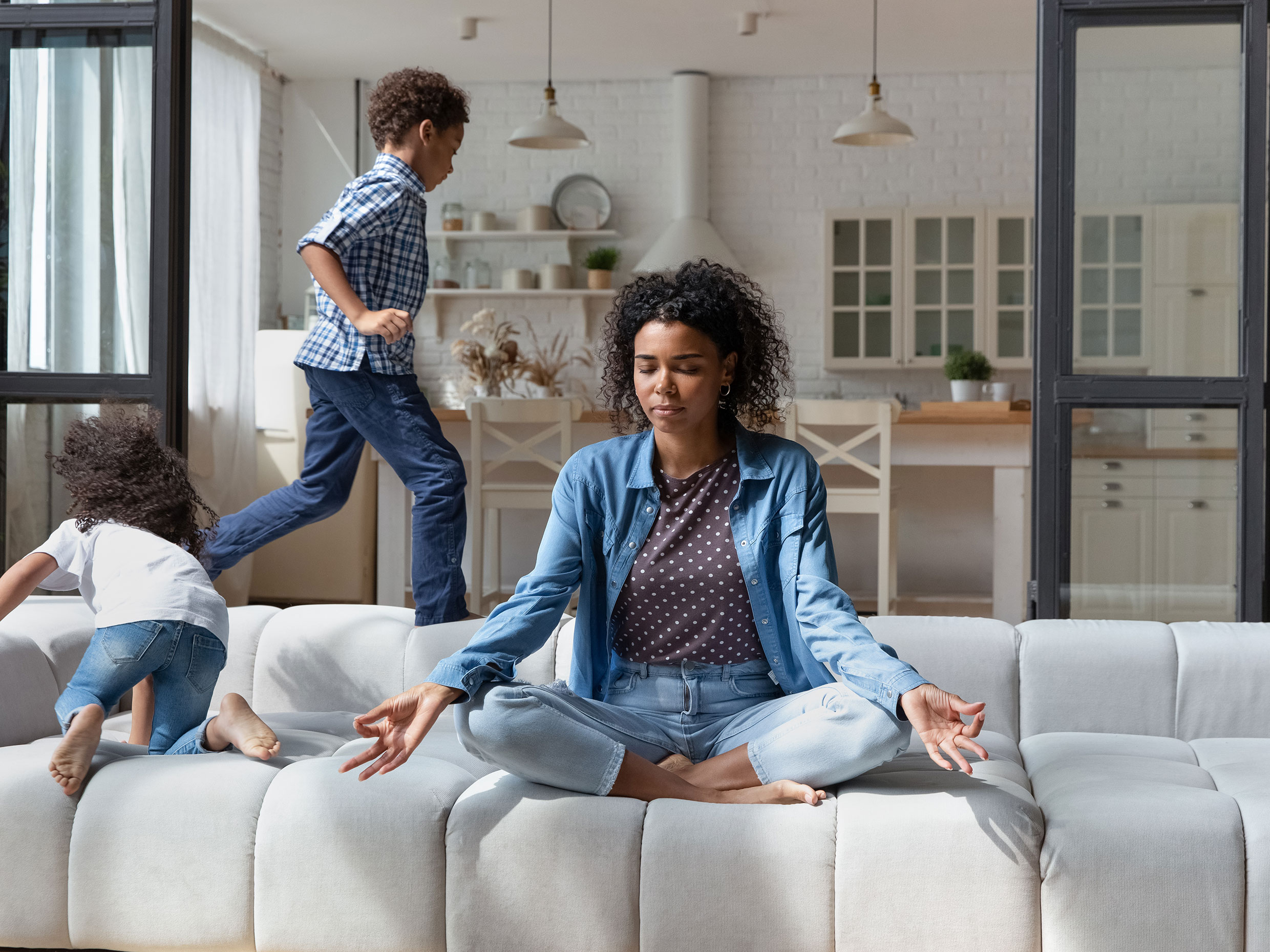 Woman meditating on couch while child runs in background.